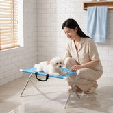 Woman grooming a small dog on a foldable pet grooming table in a bright bathroom.